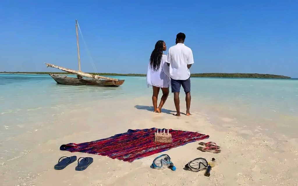 Couple standing on a sandbank beside a wooden dhow with a blanket, snorkel masks and a small cake